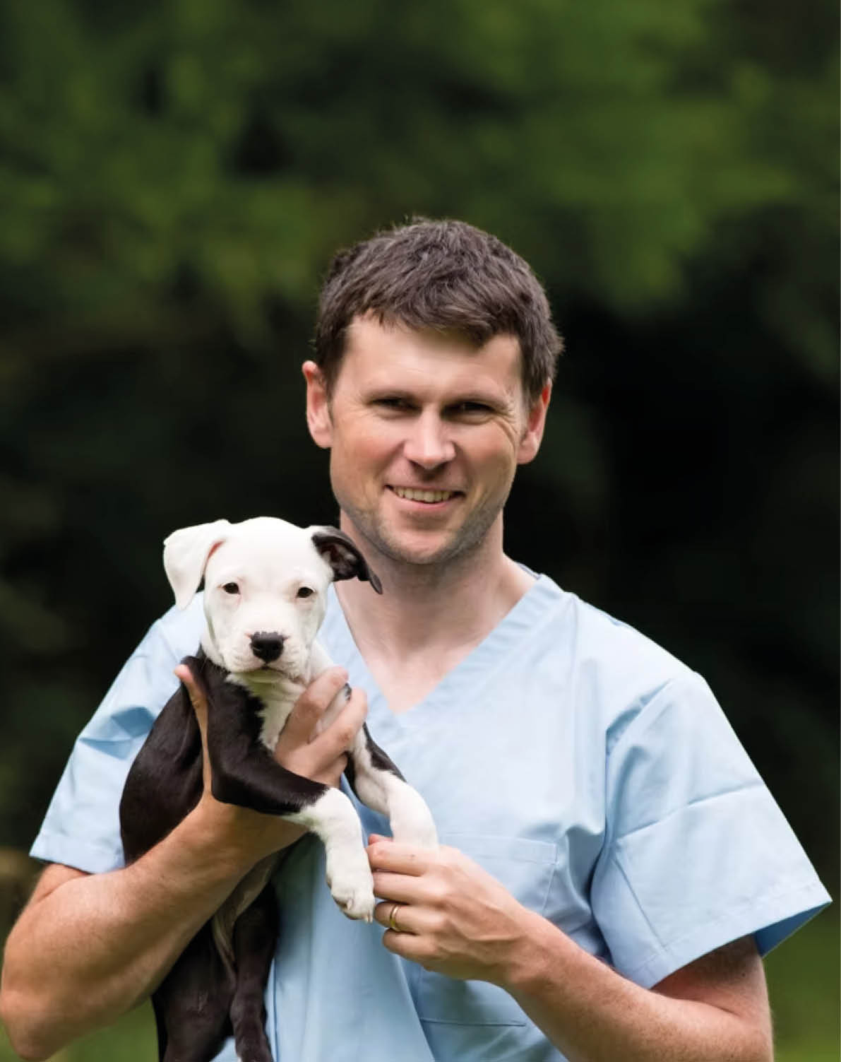 A person with short curly hair is outdoors with a blurred green background of foliage. Description generated by AI