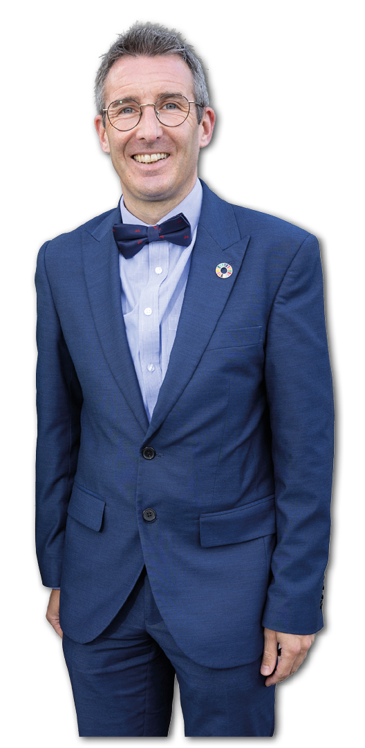 A person wearing glasses, a blue suit with a bow tie, and a circular pin on the lapel is smiling against a white background. Description generated by AI