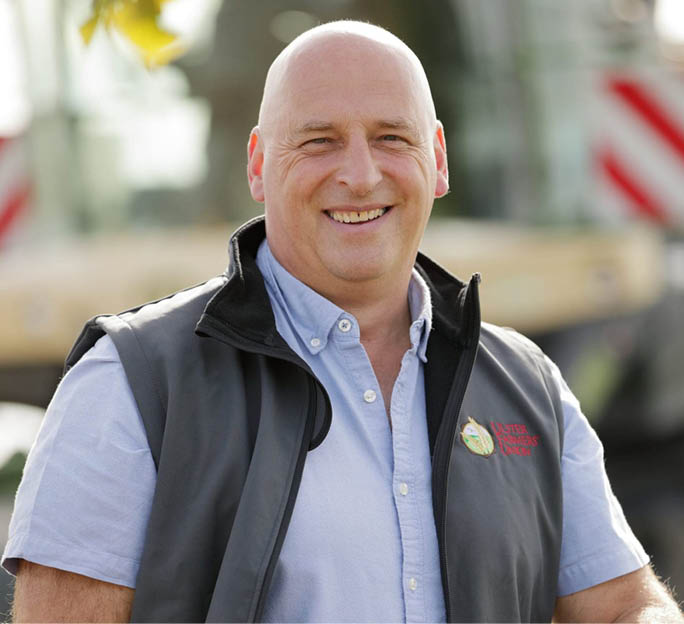 A smiling person wearing a light blue button-up shirt and a dark vest stands outdoors with a blurred background. Description generated by AI