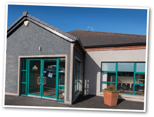 A small modern building with gray walls, teal-framed glass doors, and windows is situated under a clear blue sky with a large potted plant near the window. Description generated by AI