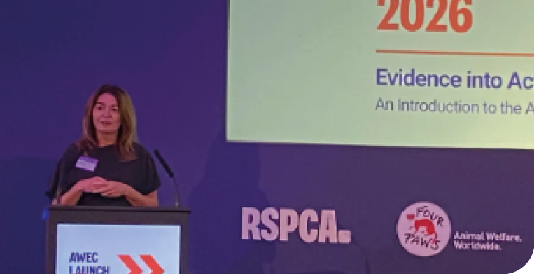 An adult with shoulder-length hair stands at a podium speaking at an event with RSPCA and animal welfare logos visible on the backdrop. Description generated by AI