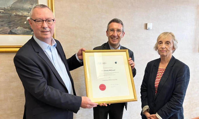 A person in a suit is holding a framed certificate awarded to Dr. Stanley McDowell for service at the Chief Executive Office of a health trust. Description generated by AI
