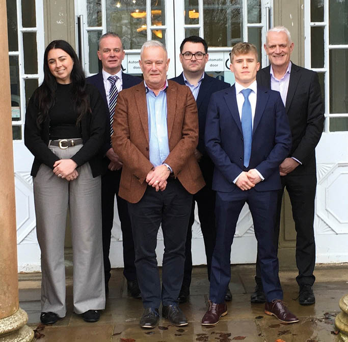 A group of five people are standing in front of a building, posing for a picture. They are all dressed in business attire, with three men wearing ties. The group consists of a man and a woman, with the woman standing slightly behind the man. They are all smiling for the camera, creating a pleasant and professional atmosphere. AI generated content