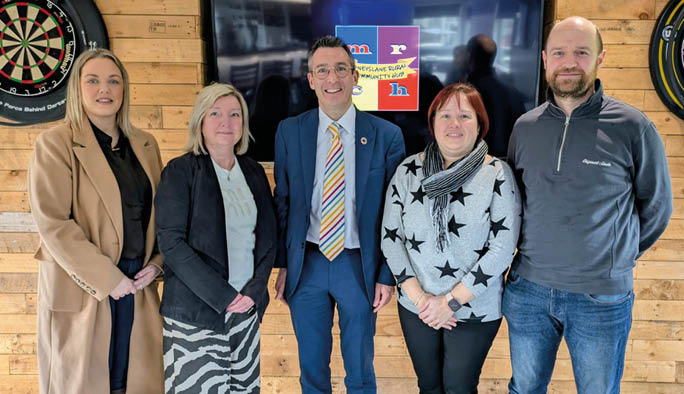 A group of five people are standing in front of a large screen TV. They are posing for a picture, with a man in a suit and tie standing in the center. The other people are standing around him, with one person on the left, one on the right, and two on the far right. AI generated content
