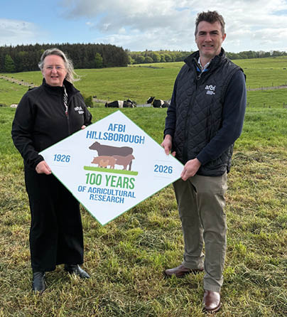 Two adults stand on grass in a rural setting holding a sign celebrating 700 years of agricultural research at AFBI Hillsborough. Description generated by AI