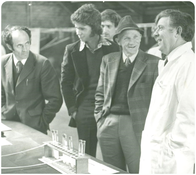 Several adults in formal and lab attire stand around a table with test tubes in a scientific or laboratory setting. Description generated by AI