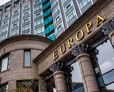 A modern multi-story building with large windows and stone columns features the word EUROPA in large gold letters on its facade. Description generated by AI