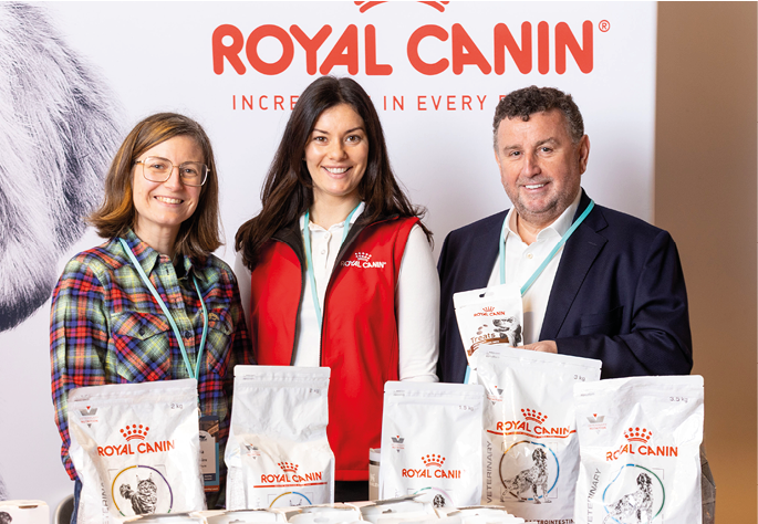 Three adults wearing event badges stand behind a table displaying various Royal Canin pet food packages with a Royal Canin banner in the background. Description generated by AI