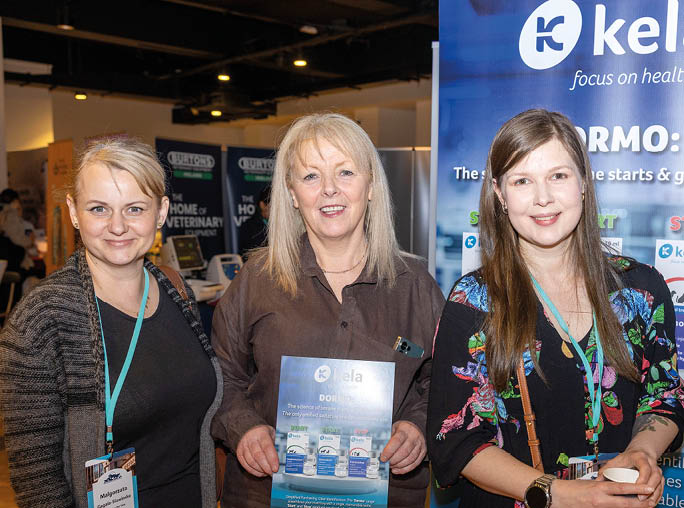 Three adults wearing lanyards stand indoors at a health-related event, with one holding a brochure and another holding a cup, in front of a display featuring the brand “kela." Description generated by AI
