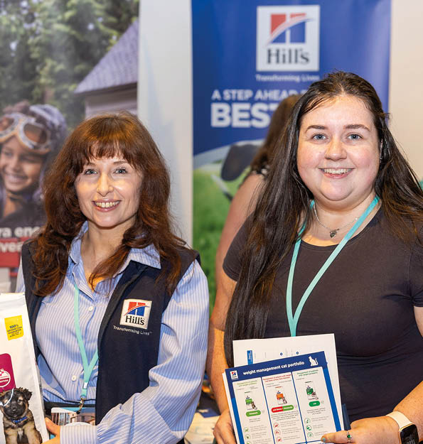 Two adults wearing lanyards stand at a promotional booth with pet food products and informational materials, with a backdrop featuring the Hill's brand. Description generated by AI