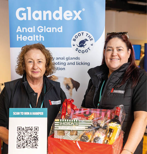Two adults wearing black shirts stand indoors holding a gift basket in front of a promotional banner for Glandex Anal Gland Health. Description generated by AI