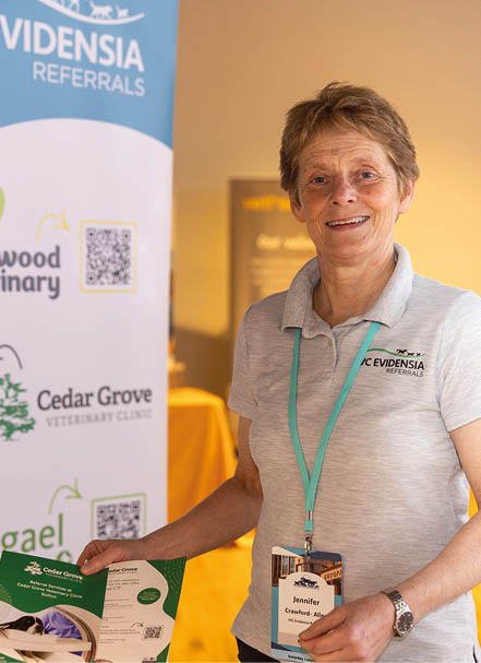 A person wearing a light gray polo shirt with a lanyard is standing indoors next to a display banner featuring veterinary clinic logos and holding informational brochures. Description generated by AI
