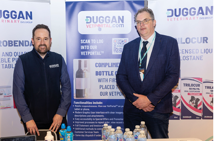 Two adults wearing business attire stand behind a table displaying various bottles and promotional materials at a veterinary product booth. Description generated by AI