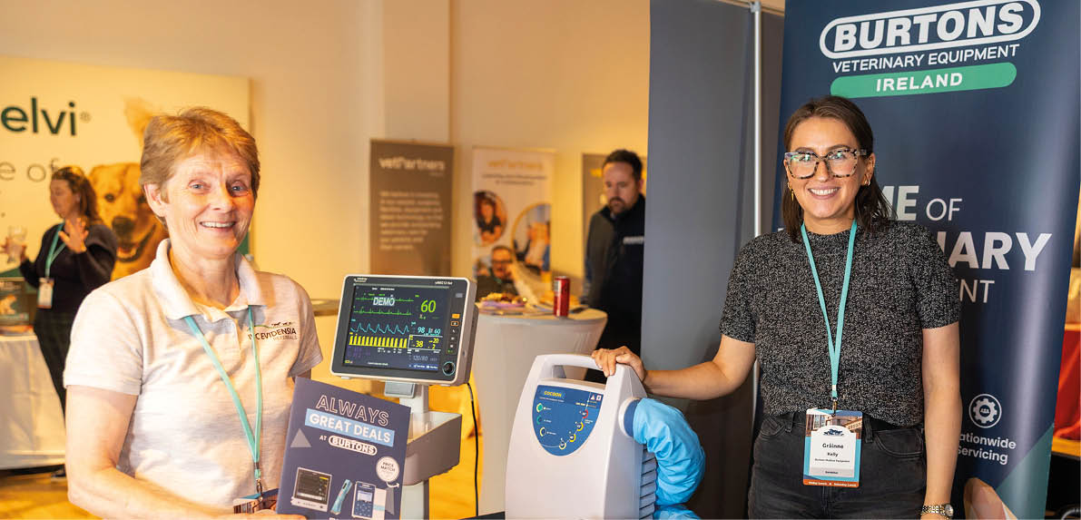 Two adults stand behind a table displaying veterinary equipment and promotional materials at an indoor event. Description generated by AI