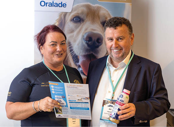Two people wearing conference badges stand in front of a large poster of a dog, with one person holding a brochure and the other holding a bottle. Description generated by AI
