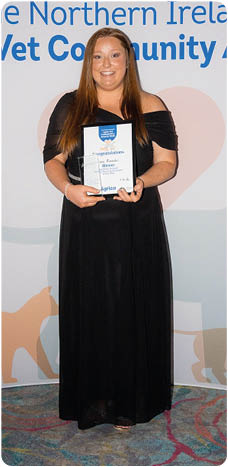 A person wearing a long black dress is standing on a colorful carpet holding an award plaque in front of a backdrop that reads The Northern Ireland Agria Vet Community Awards with animal silhouettes and sponsor logos. Description generated by AI