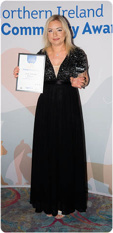 A person in a long black dress is standing on a patterned carpet holding an award certificate and a trophy in front of a backdrop with logos and text related to the Northern Ireland Agria Vet Community Awards. Description generated by AI