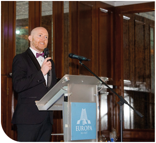 A person in formal attire with a bow tie is speaking into a microphone at a podium in a wood-paneled room. Description generated by AI