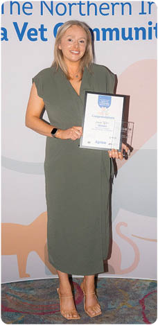 A person wearing a green dress and sandals is standing on a carpeted floor holding a framed certificate in front of a backdrop with logos and text related to the Northern Ireland Agria Vet Community Awards. Description generated by AI