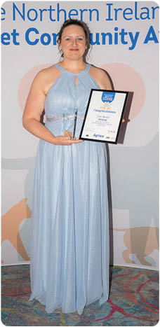 A person in a light blue formal dress is standing on a patterned carpet holding an award and a certificate in front of a backdrop for the Northern Ireland Agria Vet Community Awards. Description generated by AI