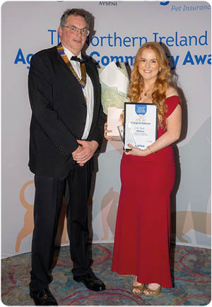 Two adults dressed formally stand in front of a backdrop with logos and text, with one holding a bouquet of flowers and the other holding a framed certificate. Description generated by AI