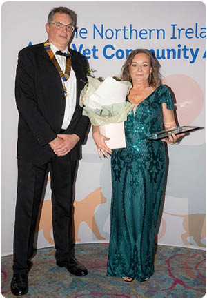 Two adults dressed formally stand in front of a backdrop with text, one holding a bouquet of flowers and a plaque. Description generated by AI