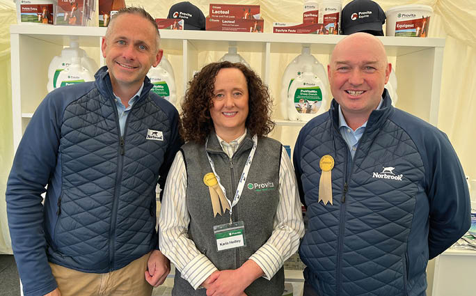 Three adults wearing branded quilted jackets and standing in front of a display shelf with various packaged products and promotional items. Description generated by AI