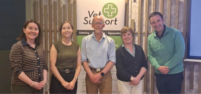Five adults stand side by side indoors in front of a Vet Support banner, with casual clothing and neutral expressions. Description generated by AI
