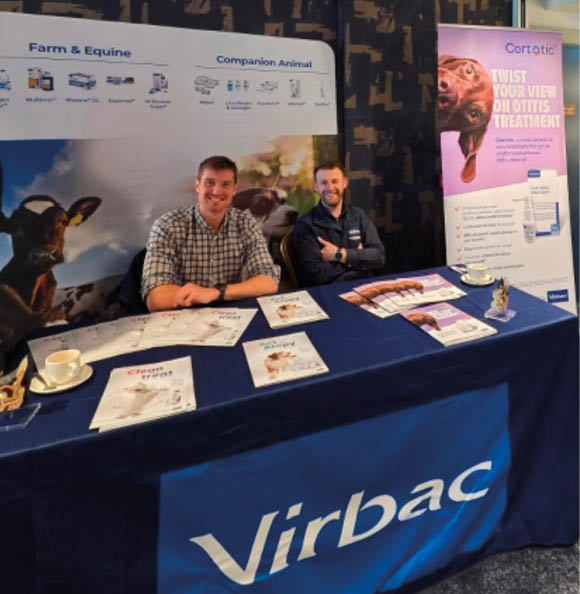 Two adults sit behind a table covered with a blue Virbac tablecloth displaying brochures and promotional materials in a carpeted indoor setting with informational banners behind them. Description generated by AI