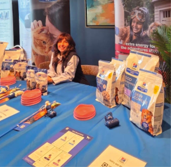 People are sitting behind a table covered with a blue cloth displaying various pet food products and promotional materials in a well-lit indoor setting. Description generated by AI