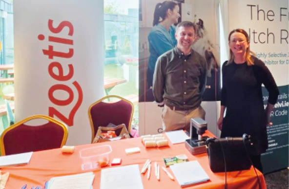 Two adults stand behind a table covered with an orange cloth displaying promotional materials and pens at a booth with banners for Zoetis and Numelvi, set in a well-lit indoor space. Description generated by AI