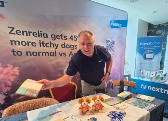 A person wearing a dark shirt is standing behind a table covered with a white cloth displaying promotional materials, two small stuffed dogs, and brochures at an indoor event with branded banners in the background. Description generated by AI