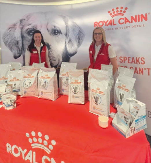 Two adults wearing red vests stand behind a table covered with a red Royal Canin tablecloth displaying various bags and containers of pet food in front of a large backdrop featuring a dog's face and the Royal Canin logo. Description generated by AI