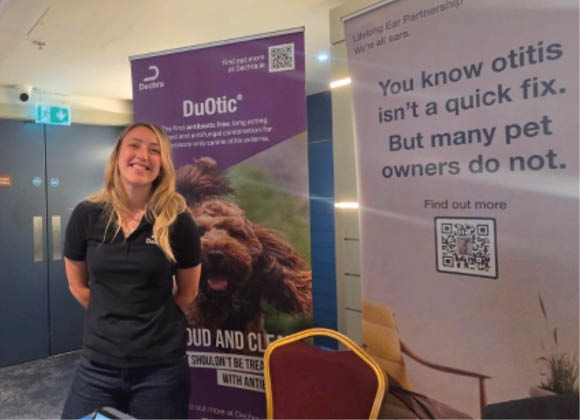 An adult with long hair stands behind a table with a laptop and promotional materials at a booth displaying banners about otitis treatment for pets. Description generated by AI