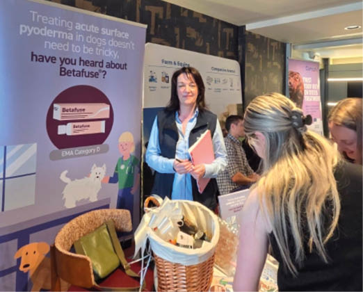 People are gathered around a table with a basket and informational materials in front of a promotional banner about treating acute surface pyoderma in dogs using Betafuse. Description generated by AI
