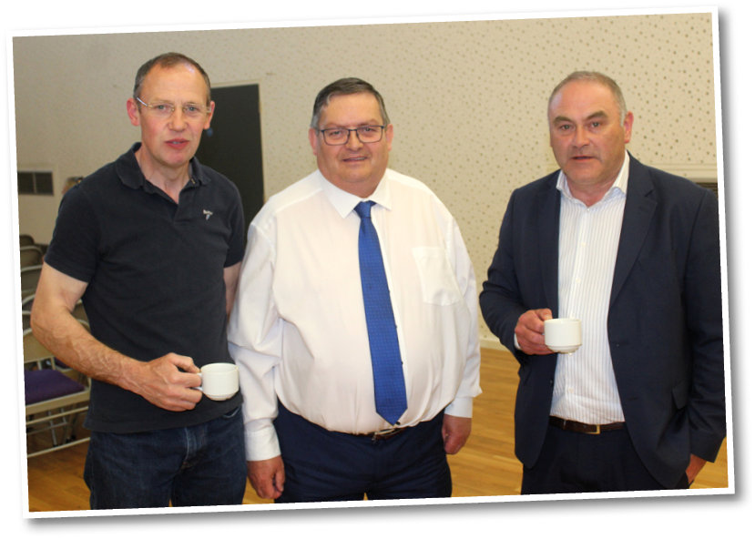 Three adults dressed in business casual attire stand indoors holding small white cups, with one wearing a white shirt and blue tie, another in a dark polo shirt, and the third in a dark suit jacket. Description generated by AI