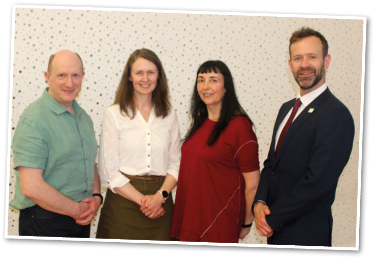 Four adults stand side by side indoors against a light-colored wall with a subtle pattern, dressed in business casual and formal attire. Description generated by AI