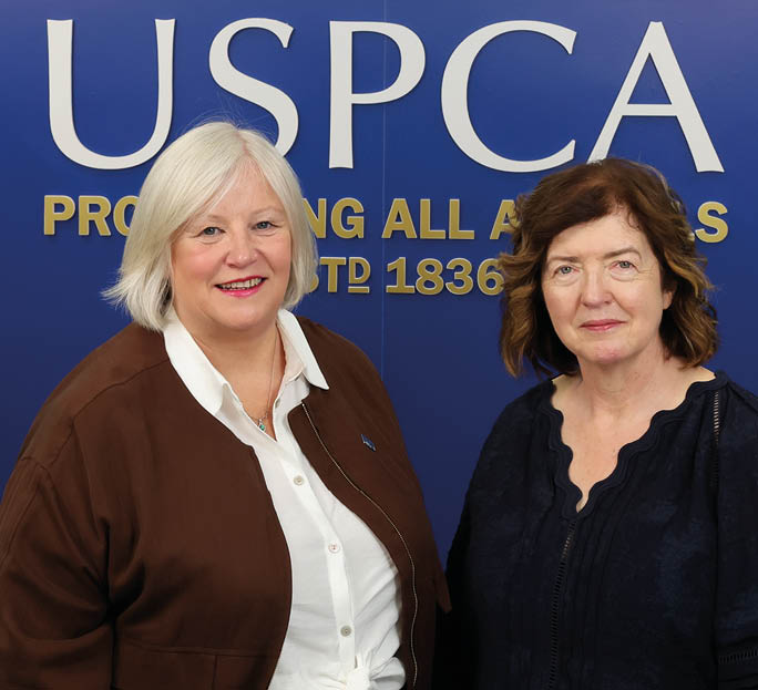 Two adults stand side by side in front of a blue backdrop with white and yellow text reading USPCA Protecting All Animals Estd 1836. Description generated by AI
