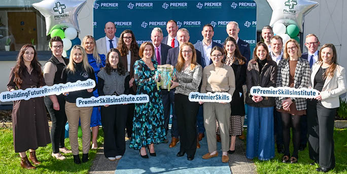 A group of adults dressed in business casual attire stand outdoors on grass and pavement holding signs with hashtags and a website, with two large star-shaped balloons and a branded backdrop behind them. Description generated by AI