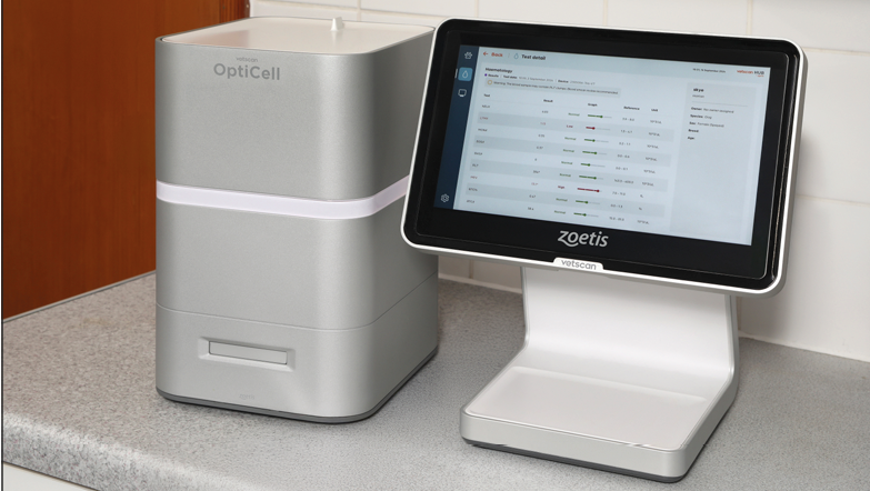 A compact silver OptiCell device is placed next to a touchscreen monitor displaying data on a gray countertop in a tiled room. Description generated by AI