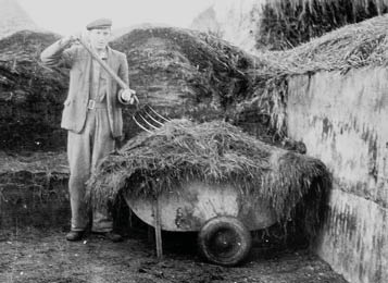 A person wearing a cap and suit stands in a large outdoor compost or manure pit holding a pitchfork with a wheelbarrow full of organic material beside them. Description generated by AI