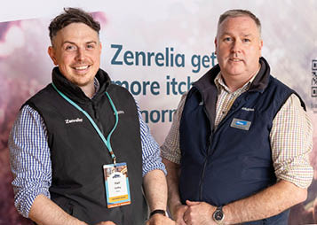 Two adults wearing casual shirts and vests stand behind a booth with promotional materials and a banner displaying the brand name Zenrelia. Description generated by AI