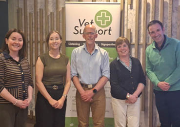 Five adults stand side by side indoors in front of a Vet Support banner, with casual clothing and neutral expressions. Description generated by AI