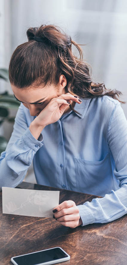 upset woman in blue blouse sitting at table with photo, smartphone and crying at home, grieving disorder concept