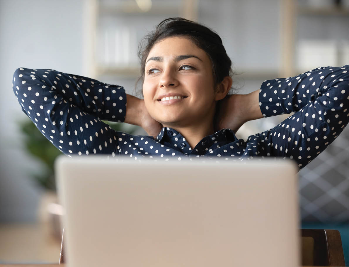 Head shot young indian woman crossed hands behind head, enjoying break time at home. Peaceful carefree millennial hindu student resting at table with computer, looking aside, dreaming of future.