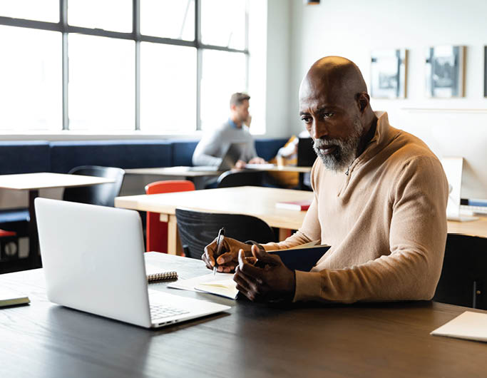 African american mature businessman writing in diary and looking at laptop in creative office. Unaltered, creative business, workplace, occupation, wireless technology, concentration.