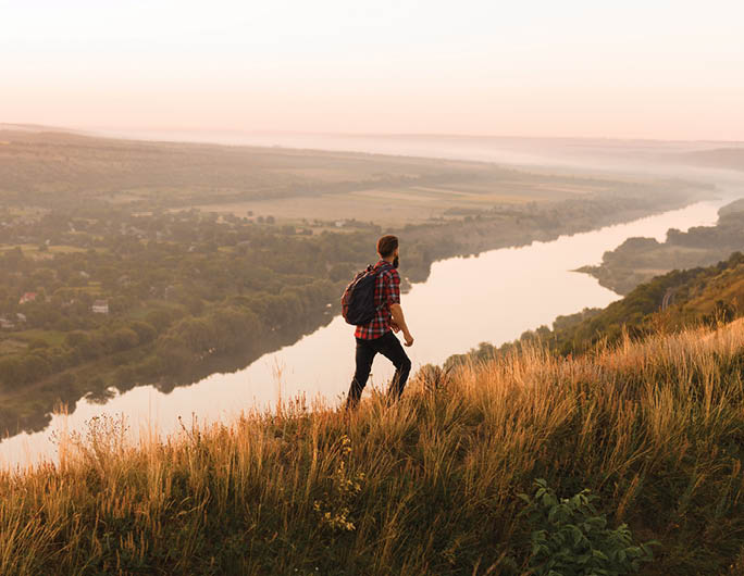 Side view of unrecognizable man with backpack admiring majestic view of river while walking in wonderful countryside during sunset