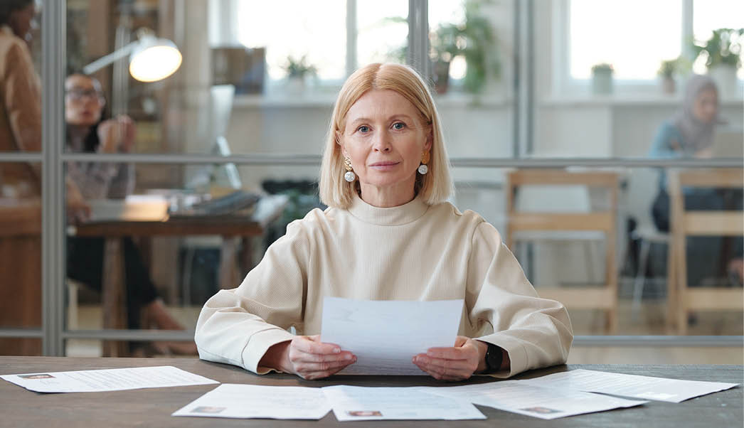 Portrait of content mature HR manager with blond hair sitting at table and reading CVs of candidates in modern open space office